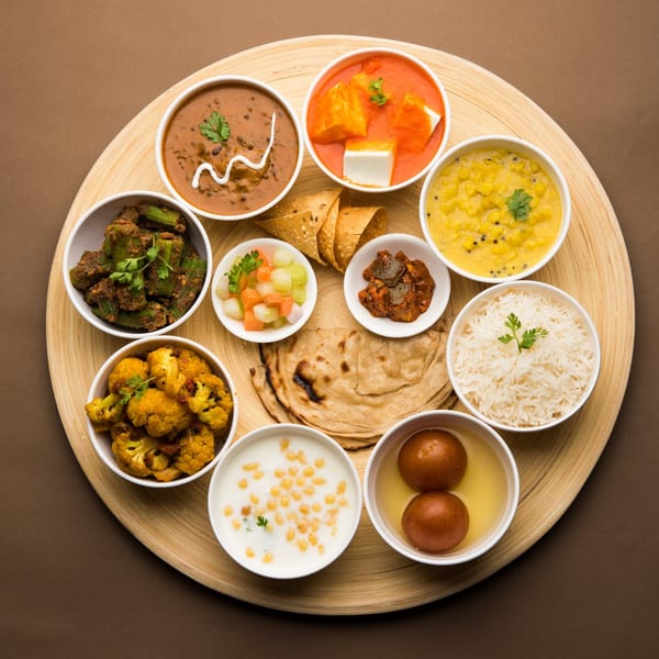 A round wooden platter holds a variety of Indian dishes, including curries, rice, flatbread, and desserts.