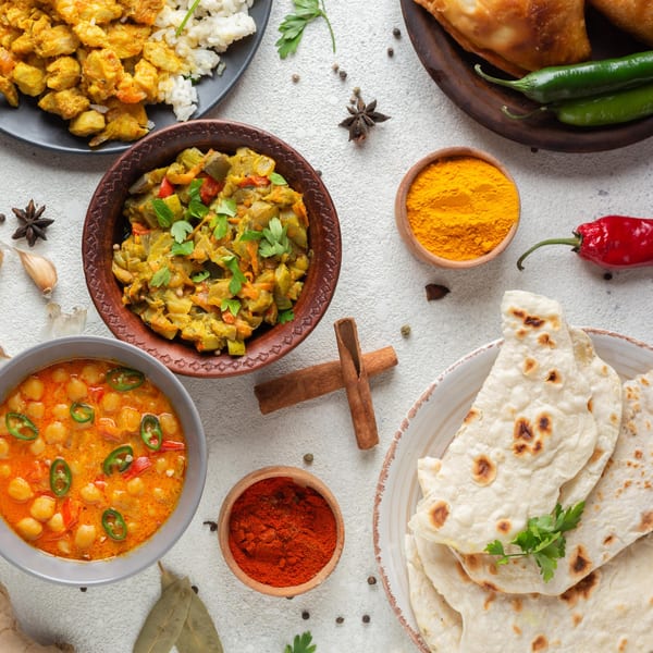 Overhead view of various Indian dishes, including curries, rice, naan, and spices on a white table.
