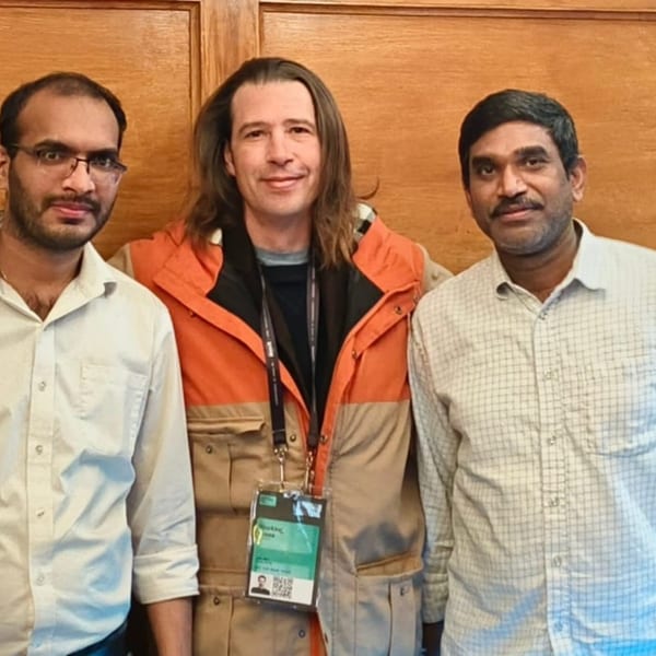 Three men, one with long hair and an orange jacket, smiling at the camera.