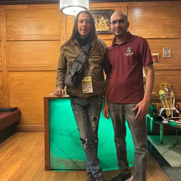 Two men standing in a restaurant, one in a maroon polo, the other in a brown jacket.