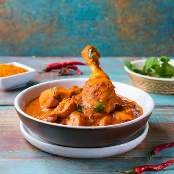 Chicken curry with drumsticks and cilantro in a bowl, with spices and herbs on a blue wooden table.