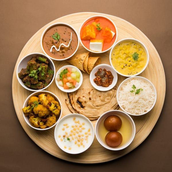 A large round wooden platter filled with various Indian dishes in white bowls, including curries, rice, bread, and desserts.