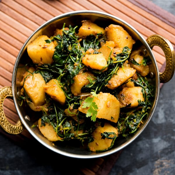 A close-up of a metal bowl filled with golden potato and green fenugreek curry.